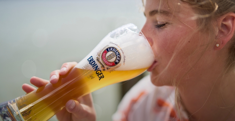 Eine Frau nimmt einen genüsslichen Schluck ERDINGER Weißbier an einem See.