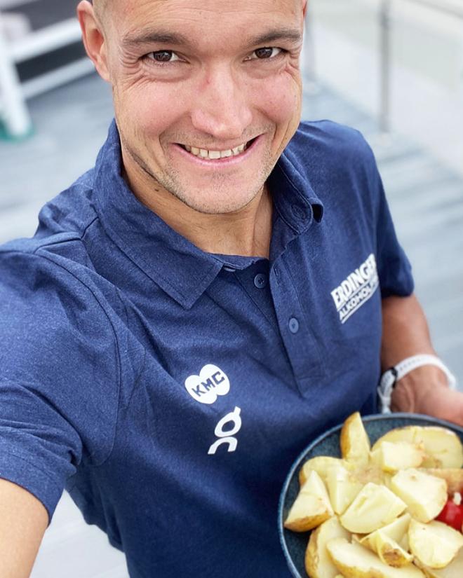 A man is smiling while holding potatoes in his hand and wearing an ERDINGER Non-Alcoholic shirt.
