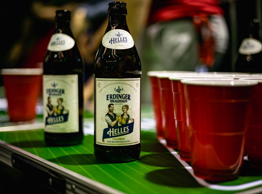 Two ERDINGER Brauhaus bottles are placed together with beer pong cups on a beer pong table.
