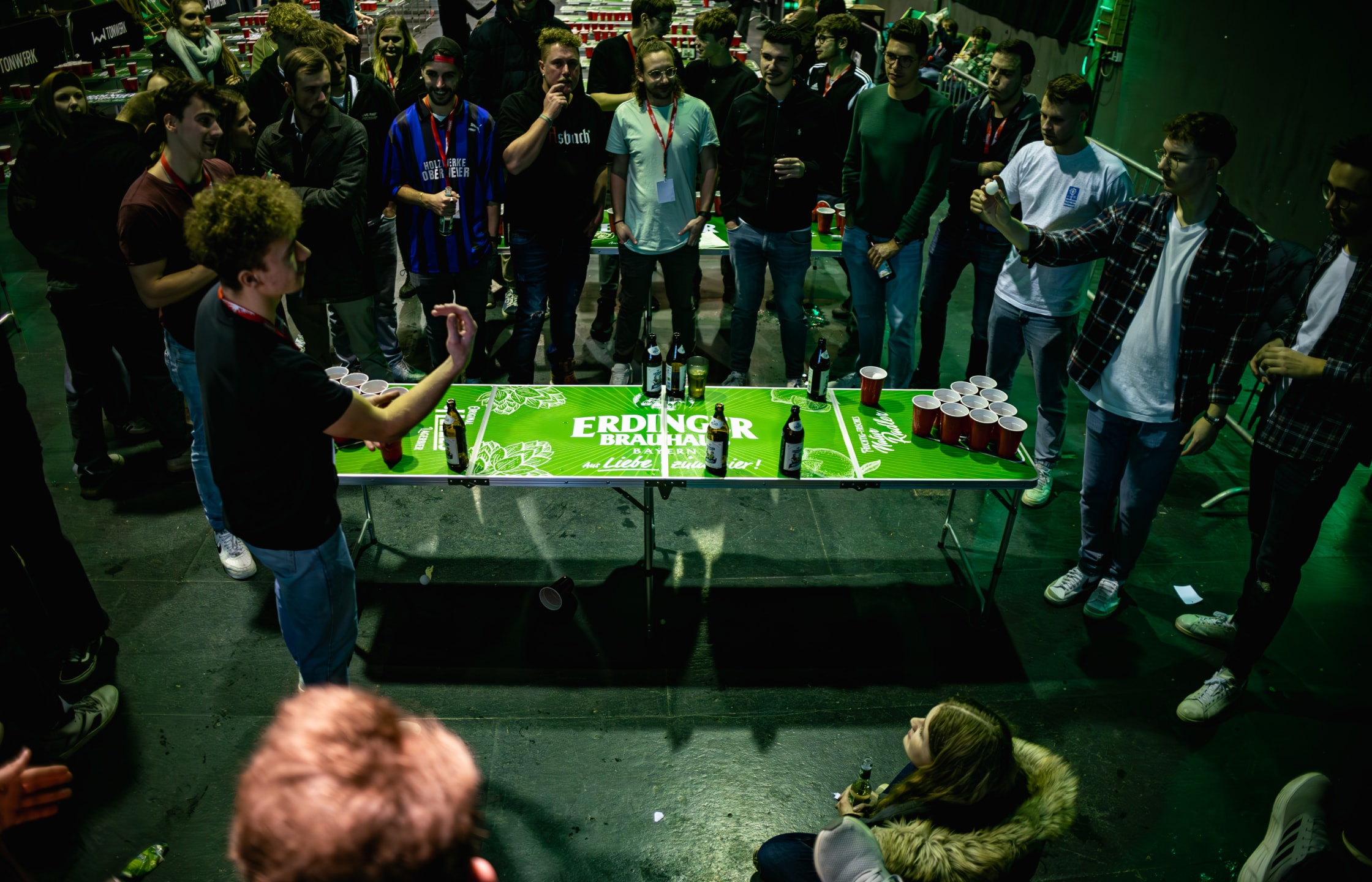 People are surrounding an ERDINGER Brauhaus beer pong table where a game is being played.