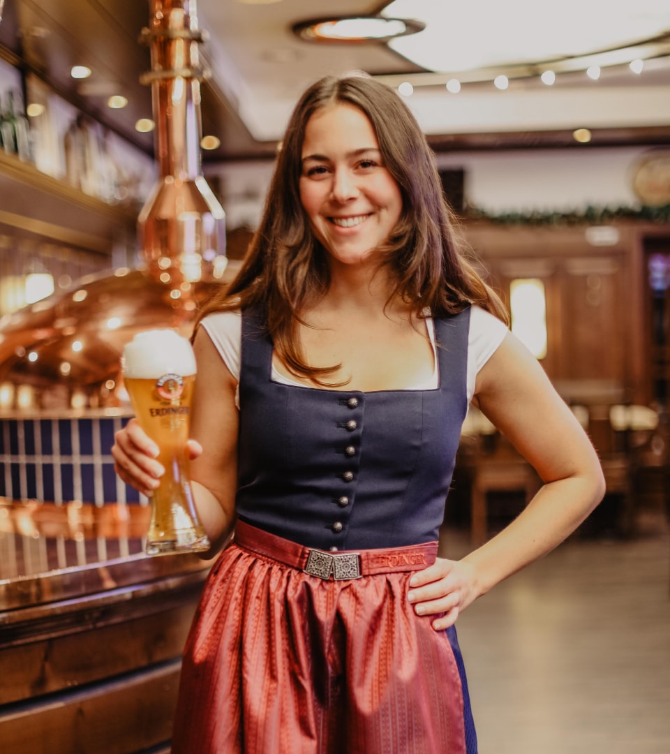 A waitress in a dirndl holds an ERDINGER Weißbier in her hand and smiles in the brewery‘s visitor center.