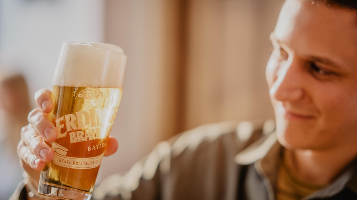 A man is holding an ERDINGER Brauhaus Hell in a glass against the light.