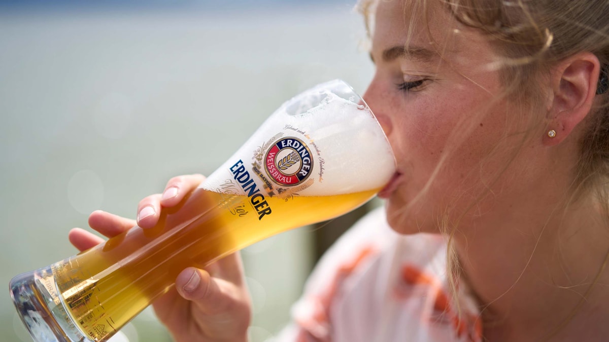 A woman takes a delightful sip of ERDINGER wheat beer by a lake.