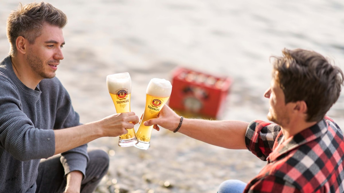 Two men toast each other with ERDINGER wheat beer by a lake.