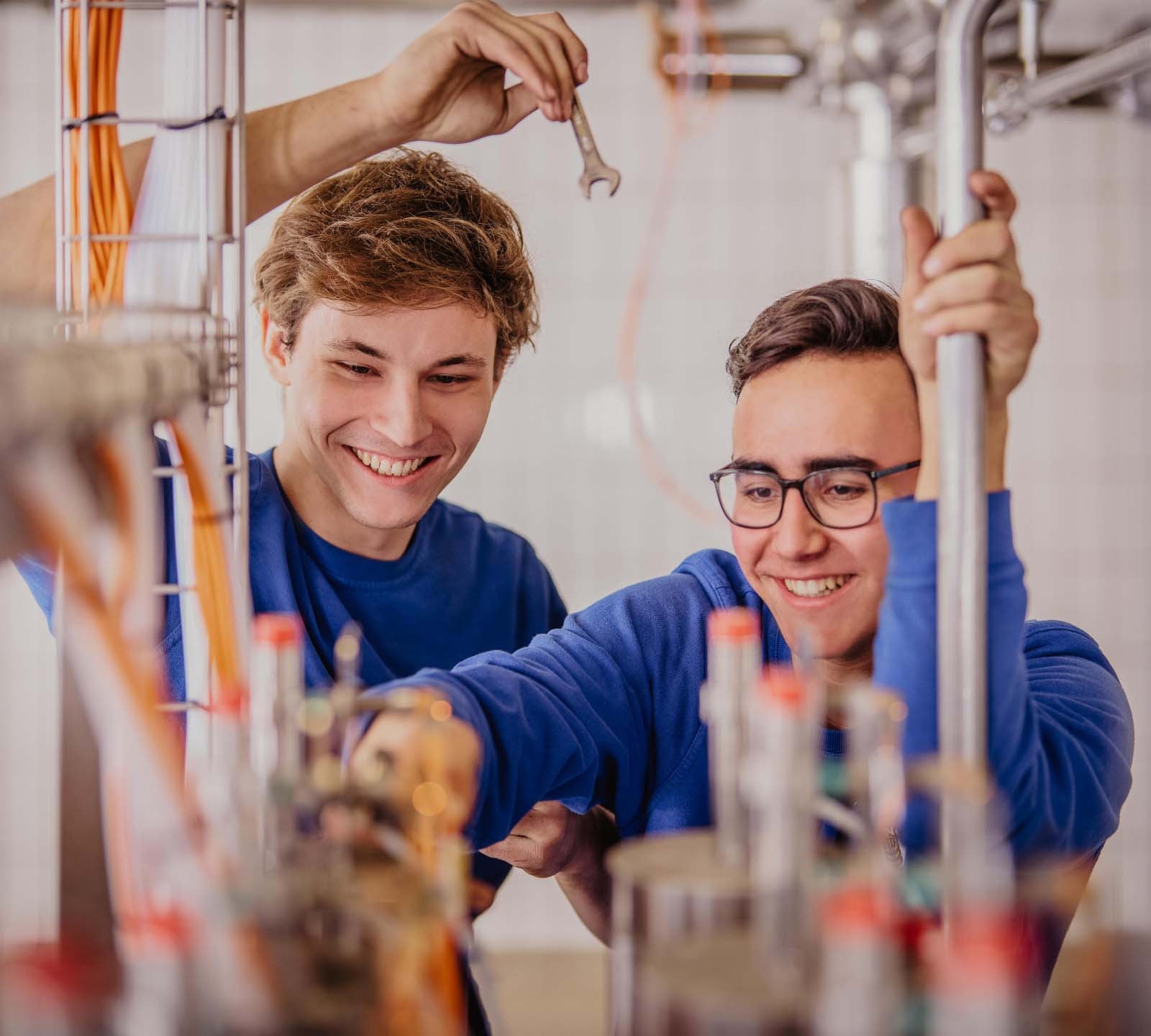 Zwei junge ERDINGER Elektroniker arbeiten zusammen und unterhalten sich lächelnd.