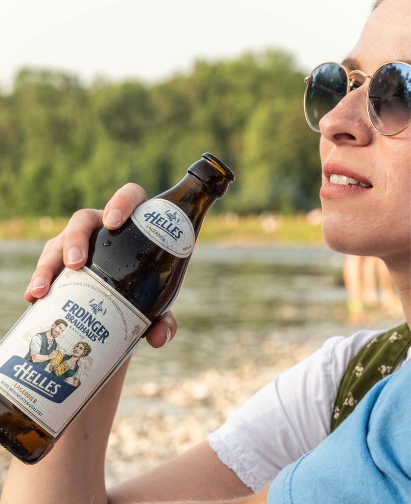 Eine Frau mit Sonnebrille sitzt an einem See mit einem ERDINGER Brauhaus Hell.