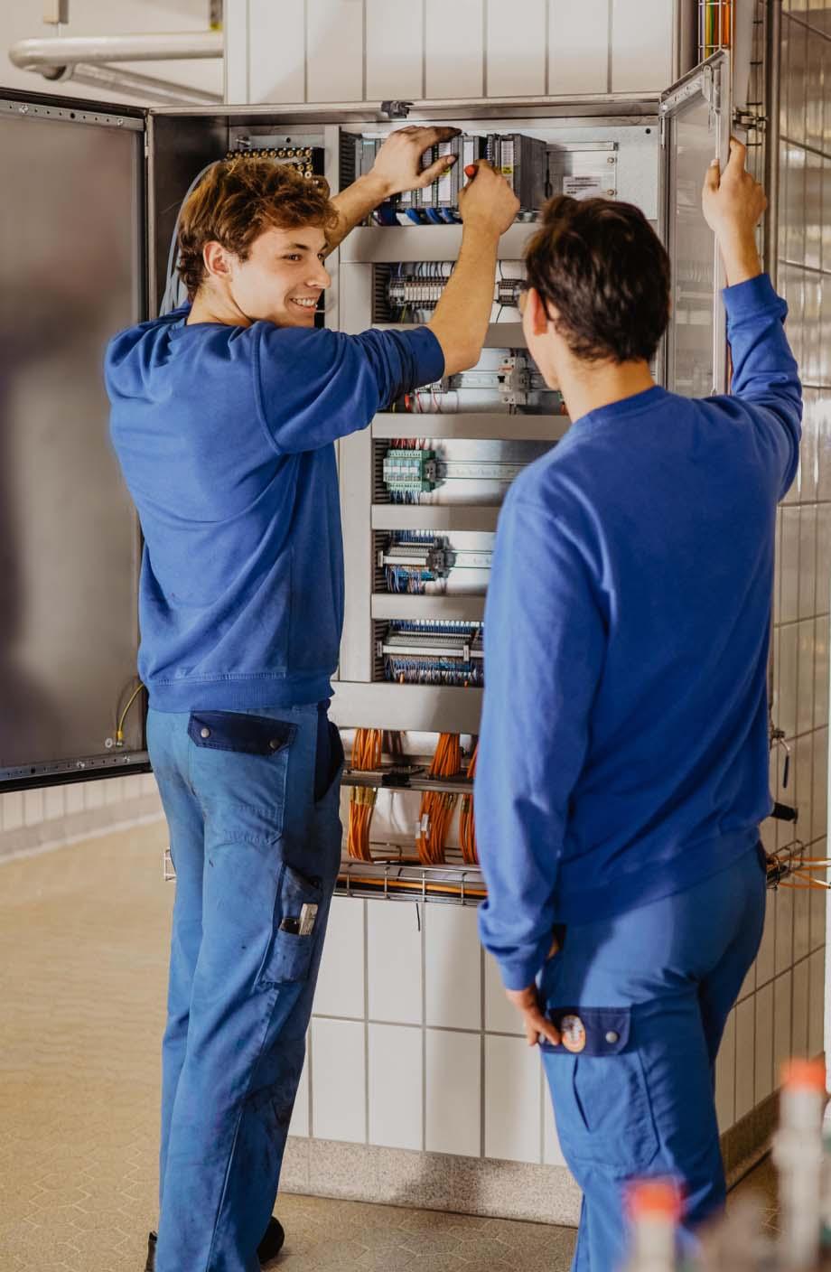 Zwei junge ERDINGER Elektroniker arbeiten zusammen an einem Schaltkasten.