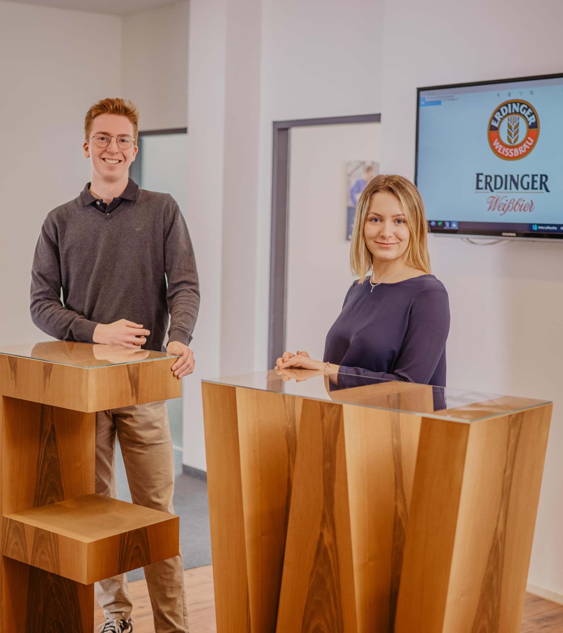 Zwei ERDINGER auszubildende Industriekaufleute stehend lächelnd im Empfangsbereich.